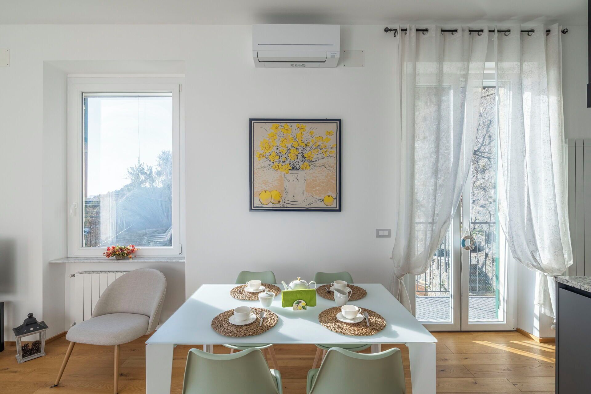 Een eetruimte met een witte tafel gedekt voor vier personen, omringd door stoelen. Tegen de muur staat een radiator, er is een raam met uitzicht op bomen en er hangt een kunstwerk boven de tafel.
