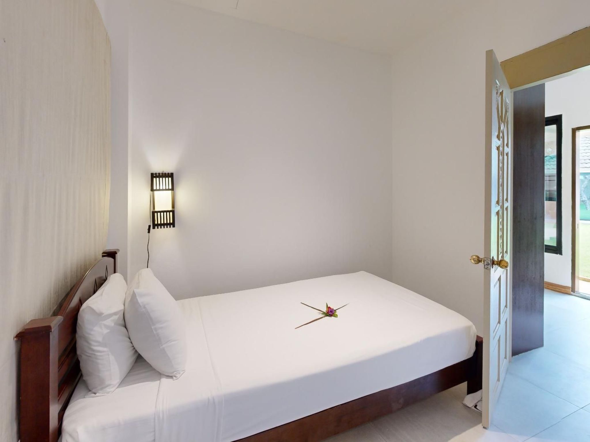A single-bed room with a wooden headboard, white bedding, a wall-mounted light fixture, and a door leading to a balcony.