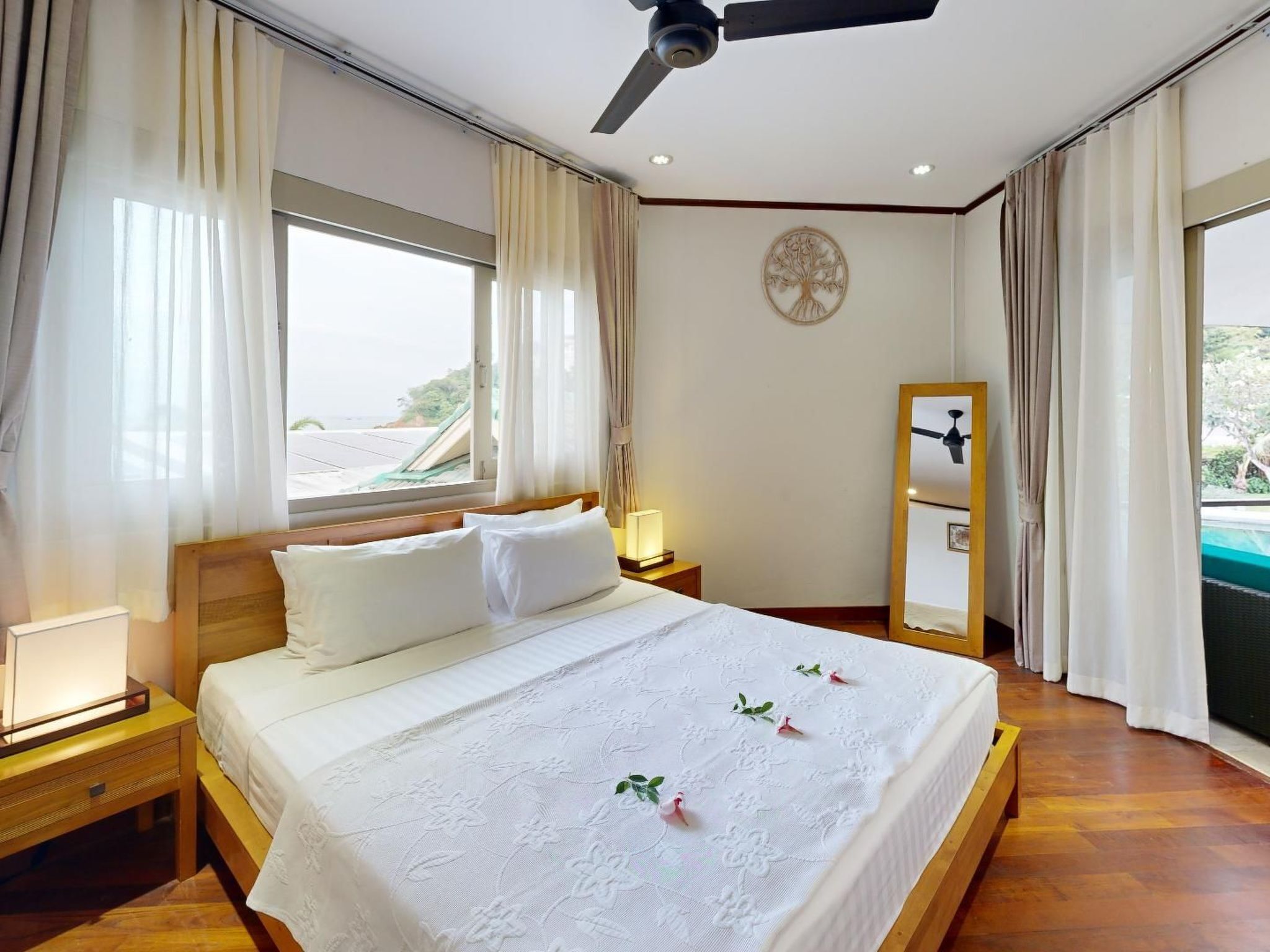Ein Hotelzimmer mit einem großen Bett, einem Deckenventilator, einem Nachttisch mit Lampe und Blick aufs Meer durch das Fenster.