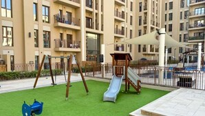 Un'area giochi con altalene, uno scivolo e una struttura per arrampicarsi, situata in una zona residenziale con palazzi condominiali sullo sfondo.