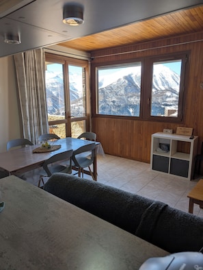 Une pièce chaleureuse avec un plafond en bois, une table à manger avec des chaises, un canapé et une vue sur des montagnes enneigées, visible à travers de grandes fenêtres.