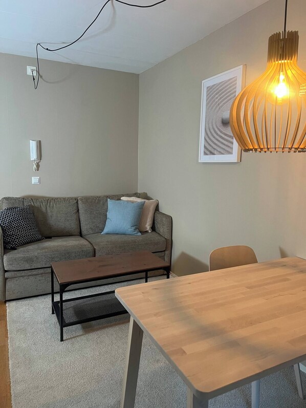 A modern living room with a sectional sofa, a wooden coffee table, a dining table, a hanging pendant light, and a framed artwork on the wall.