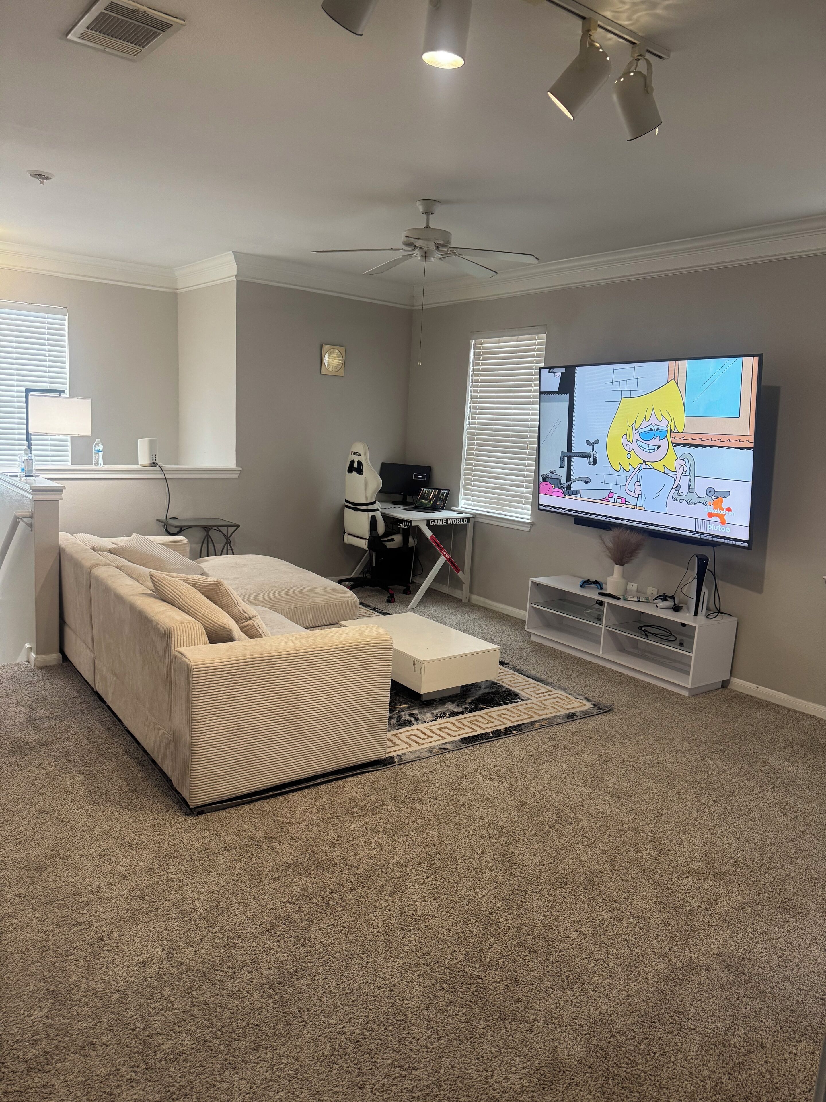 A living room with a beige sofa, a TV, a gaming chair, and a desk with a computer.