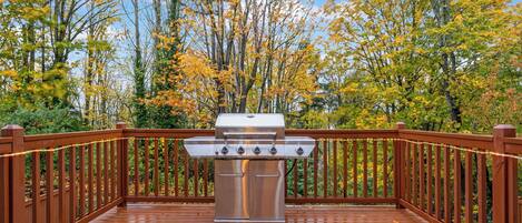 Una parrilla de barbacoa en una terraza de madera con barandilla, rodeada de árboles con follaje otoñal.