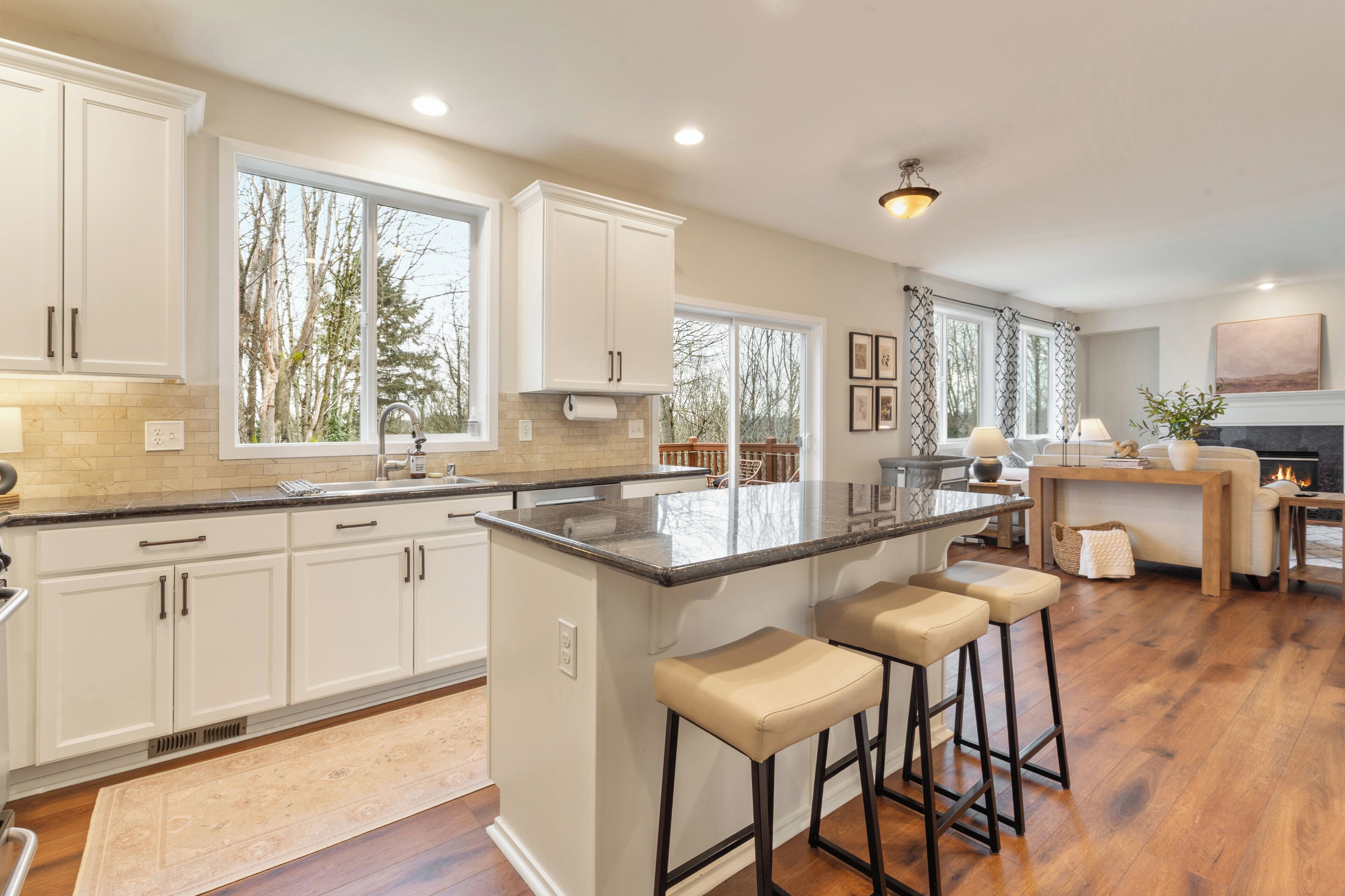 A spacious kitchen with white cabinets, a large island with stools, and a view of trees through the windows.