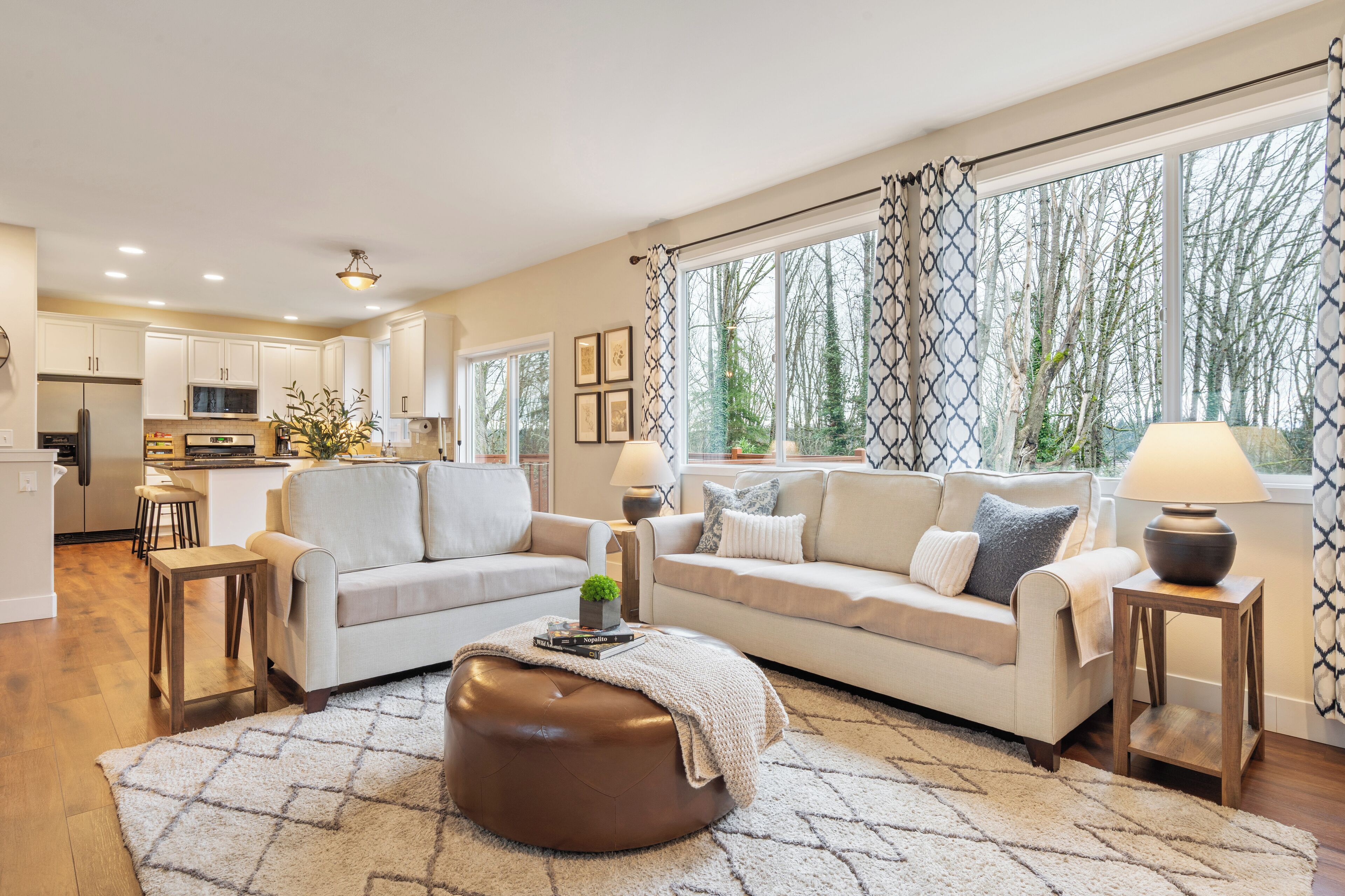 A modern living room with a sofa, ottoman, and coffee table, connected to a kitchen area with white cabinets and a dining space.