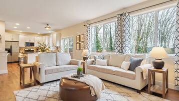 A modern living room with a sofa, ottoman, and coffee table, connected to a kitchen area with white cabinets and a dining space.