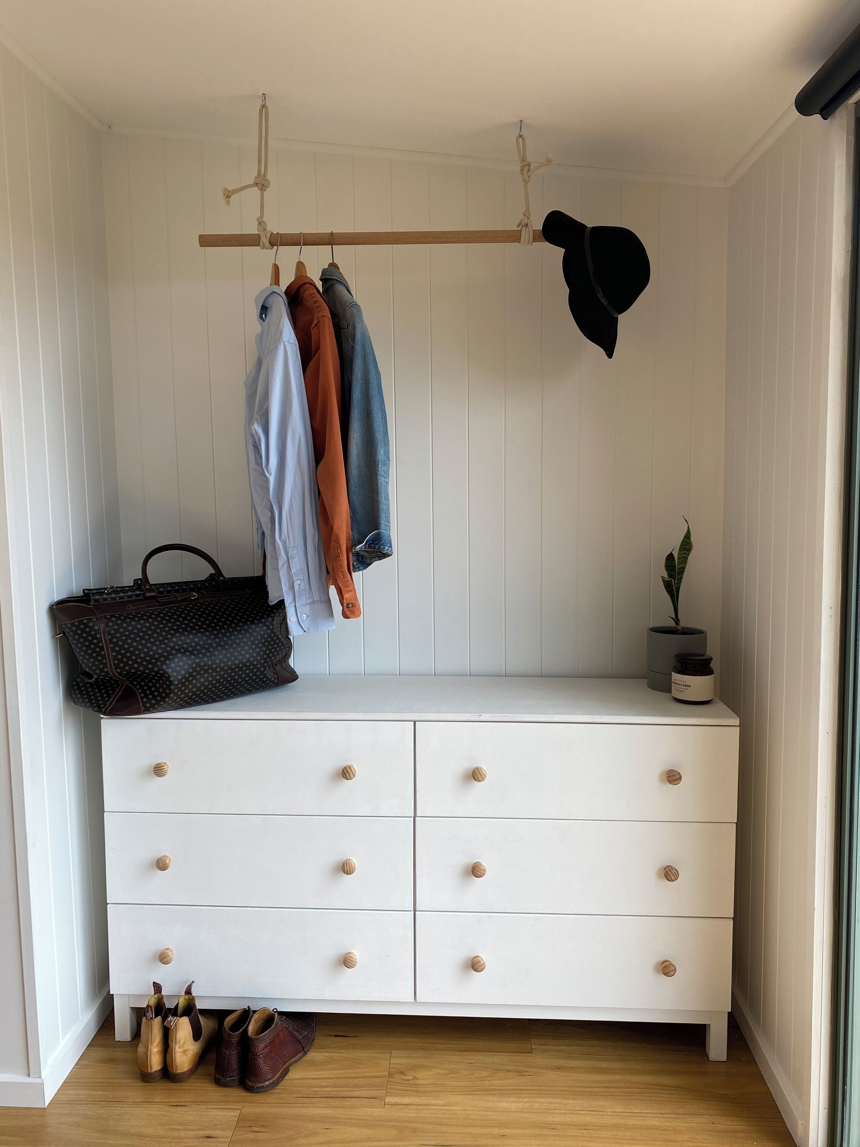 A white dresser with wooden knobs, a hanging coat rack with clothes, a black bag, a potted plant, and a pair of shoes on the floor.
