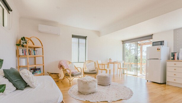 A spacious living area with a sofa, ottomans, a wooden bookshelf, a dining table, and a refrigerator. - Secluded bush get away in Geelong - only a short drive to CBD and beaches. (Belmont)