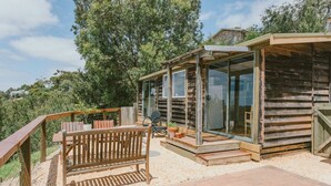 A rustic wooden cabin with a glass-enclosed porch, a wooden bench, and a small table with potted plants. - Secluded bush get away in Geelong - only a short drive to CBD and beaches. (Belmont)