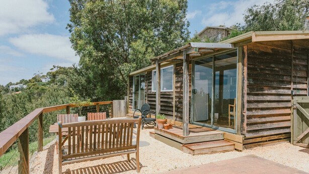 Une cabane rustique en bois, dotée d’un porche vitré, d’un banc en bois et d’une petite table agrémentée de plantes en pot.