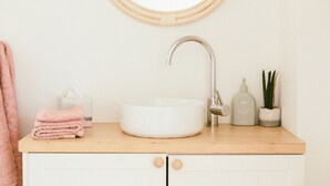 Une salle de bain avec un miroir rond, un lavabo blanc, un plan de toilette en bois, une serviette, une boîte de mouchoirs et une plante.