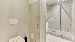 A modern bathroom with beige tiles, a glass shower enclosure, a toilet with two rolls of toilet paper, and a black cleaning brush.