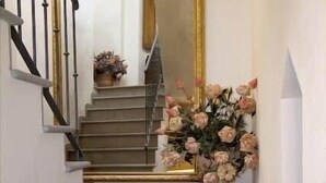 Una escalera con barandilla negra, un espejo con marco dorado y un jarrón con flores en el descansillo.