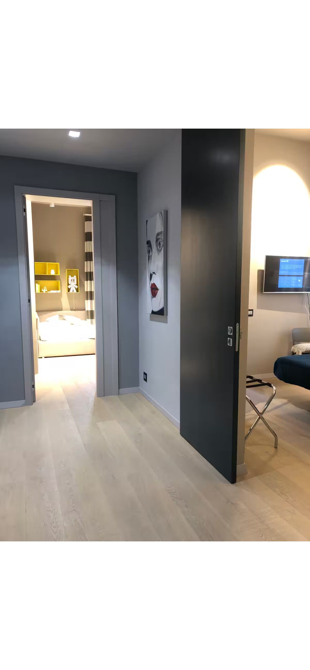 A modern room with a wooden floor, a dark door, a wall-mounted TV, and a bed in the background.
