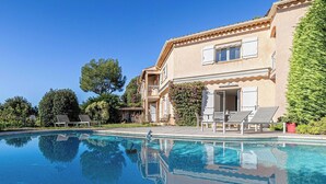 A poolside view of a two-story house with a clear pool, outdoor seating, and greenery. - Quiet House with Pool (Biot)