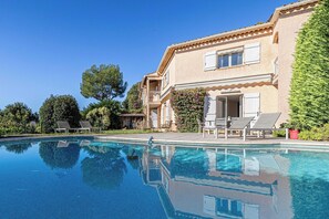 Blick auf einen Pool vor einem zweistöckigen Haus mit Sitzgelegenheiten im Freien und viel Grün.