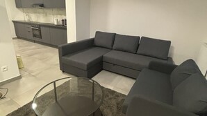 Un salon moderne avec un canapé d’angle, une table basse en verre et une cuisine en arrière-plan.