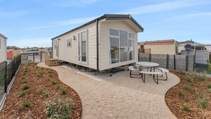 Une maison mobile dotée d’un patio, comprenant une table et des chaises, le tout entouré d’une cour clôturée avec du paillis et de petites plantes.