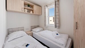 A compact hotel room with two single beds, a wooden wardrobe, a small bedside table, a window with curtains, and a view of the outside.