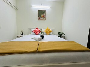 Una cama bien hecha con una manta blanca y amarilla, dos almohadas con diseños geométricos y una pequeña planta en maceta sobre la mesita de noche.