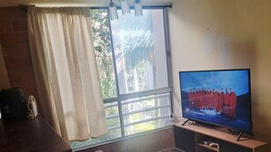 Ein Wohnzimmer mit einer Couch, einem Fernseher auf einem Holzständer, einem Fenster mit Vorhängen und einer Deckenleuchte.