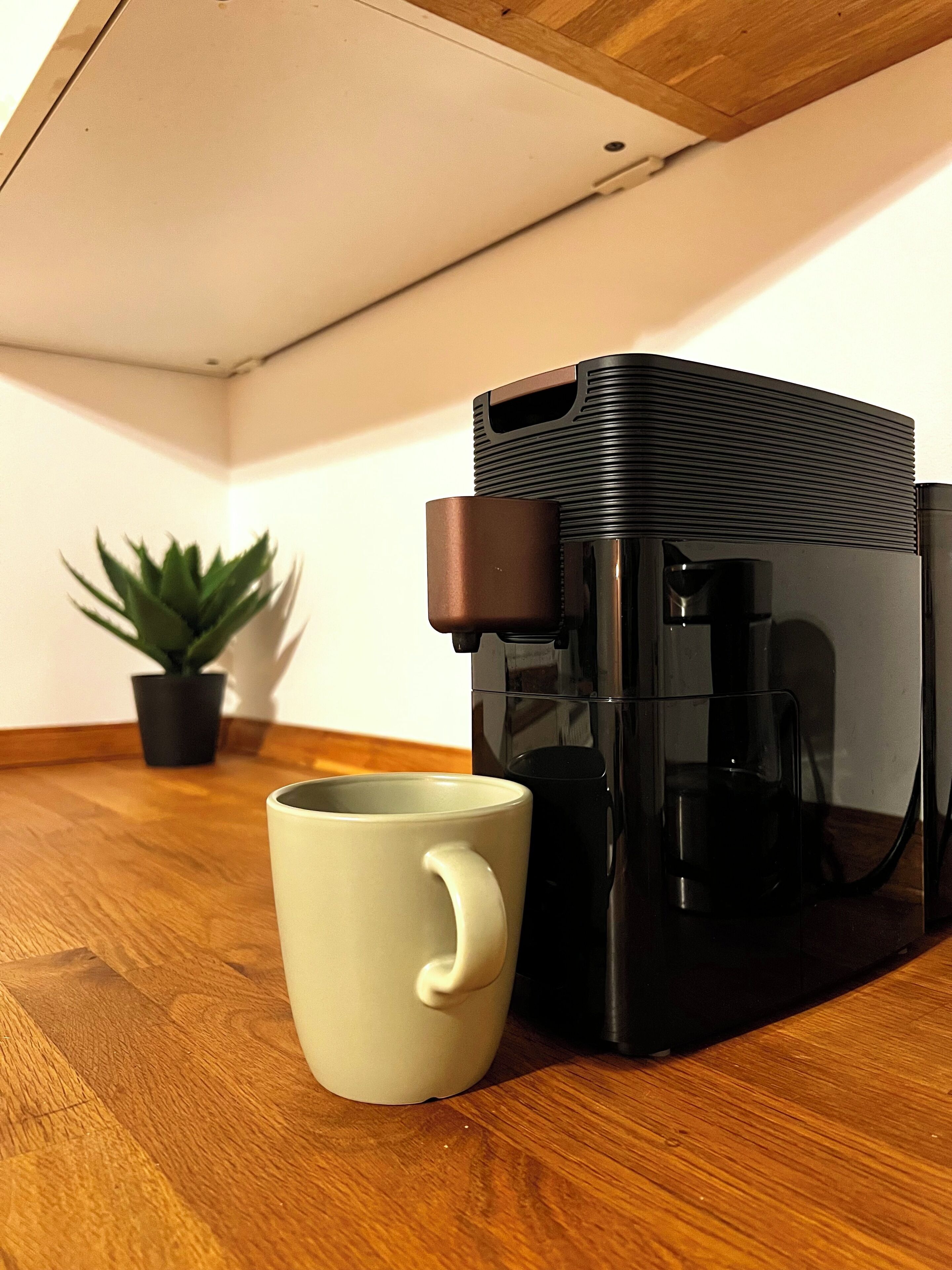 A coffee maker on a wooden table with a white mug beside it.