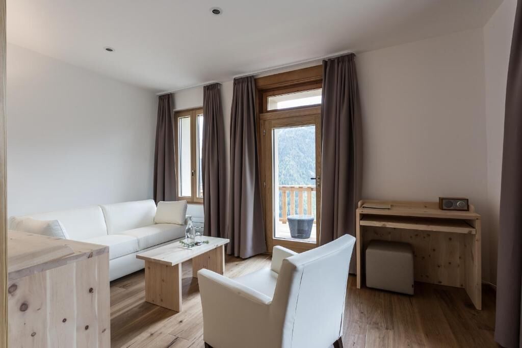 Una sala de estar moderna con un sofá blanco, una mesa de centro de madera, un sillón blanco y un escritorio de madera con una radio. Amplias ventanas con cortinas ofrecen vistas al exterior.