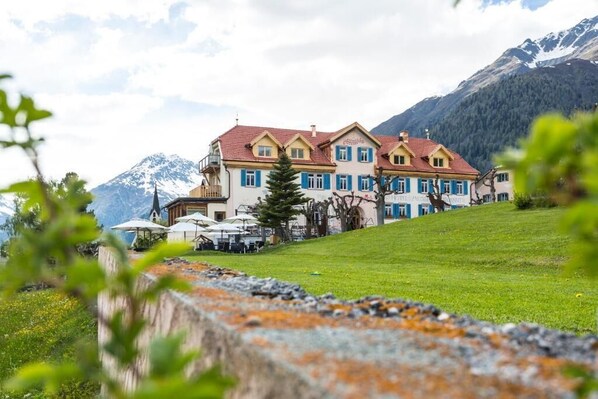 A large, multi-story building with a red roof and blue shutters, situated on a grassy slope with mountains in the background. - Meisser Hotel "superior" (Guarda)
