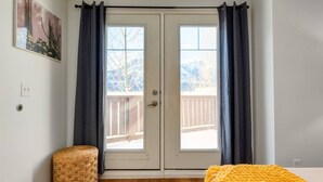 Una habitación con suelo de madera, una silla de mimbre, una manta amarilla y una puerta que da a un balcón con vistas a árboles y montañas.