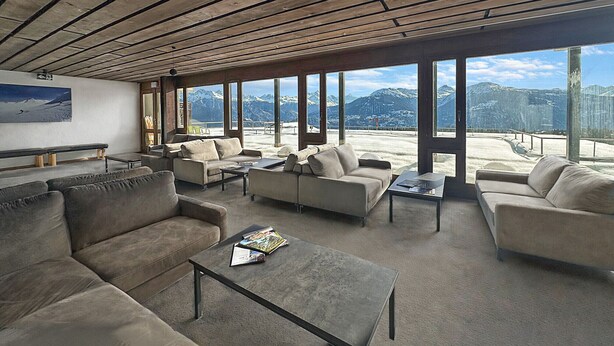 Amplia sala de estar con varios sofás y una mesa de centro, ventanas grandes que ofrecen vistas a las montañas y techo de madera.