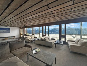 Ein großzügiger Wohnbereich mit mehreren Sofas und einem Couchtisch, große Fenster mit Blick auf die Berge und eine Holzbalkendecke.