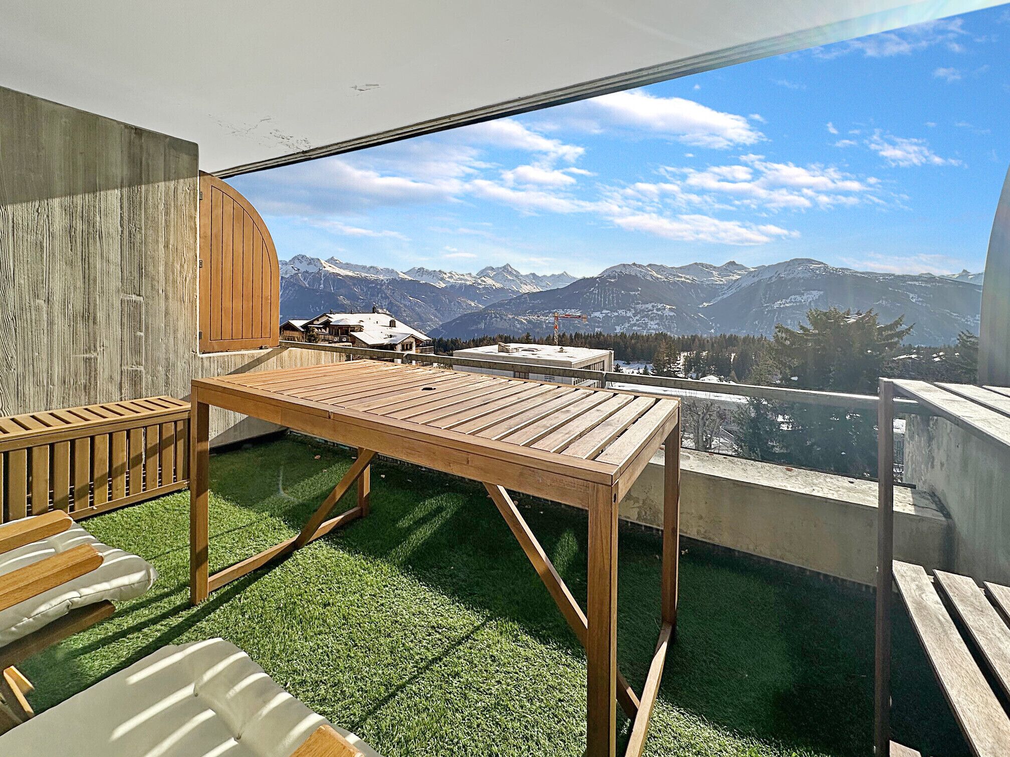 Un balcon avec une table et des chaises en bois, une vue sur des montagnes enneigées et un ciel dégagé.