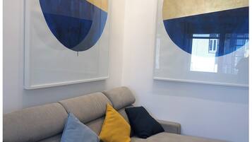 Ein modernes Wohnzimmer mit einem grauen Ecksofa, einem kleinen Hocker und einem Couchtisch. Drei Kissen in Blau, Gelb und Schwarz sind vorhanden. Zwei große abstrakte Gemälde mit Gold- und Blautönen hängen an der Wand.