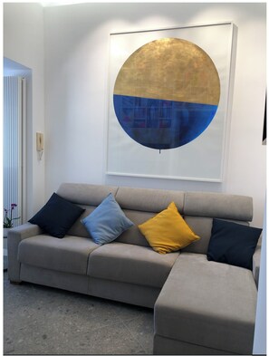 Un salon avec un canapé beige, trois coussins aux couleurs bleu, jaune et bleu foncé, et un grand tableau abstrait accroché au mur.