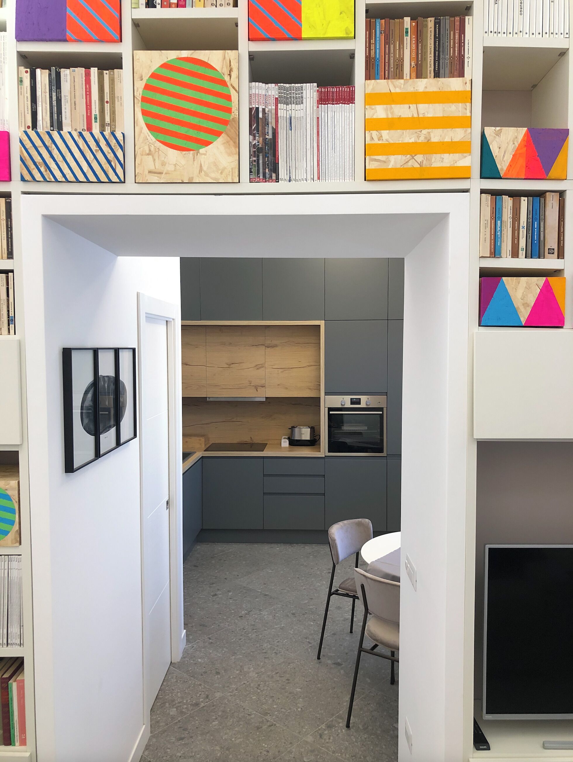 Une cuisine compacte dotée d’un four encastré, d’un petit coin repas avec une chaise, et d’une bibliothèque contenant divers livres et objets décoratifs.