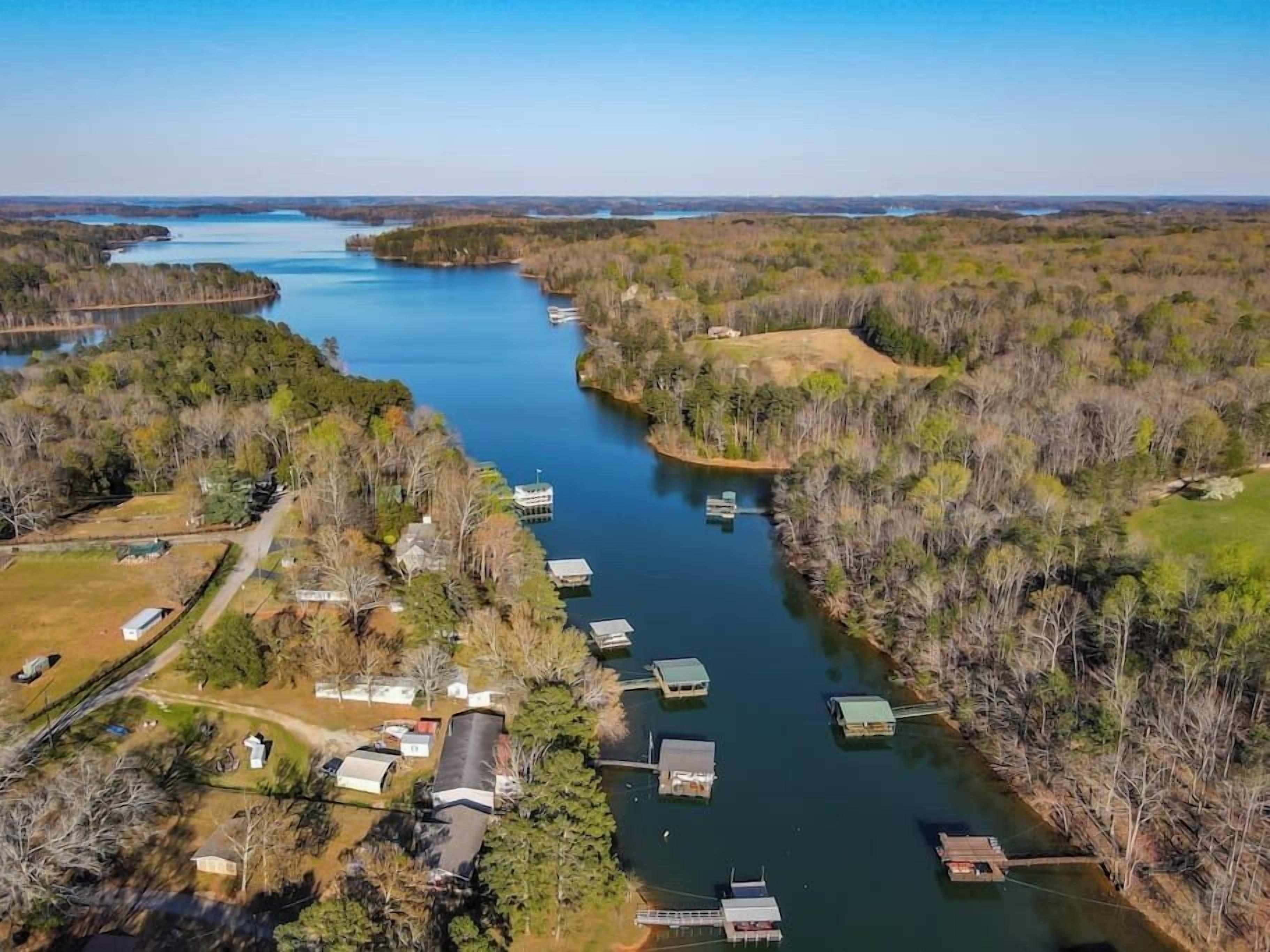An aerial view of a riverside property with several docked boats, surrounded by dense forest and a few buildings.