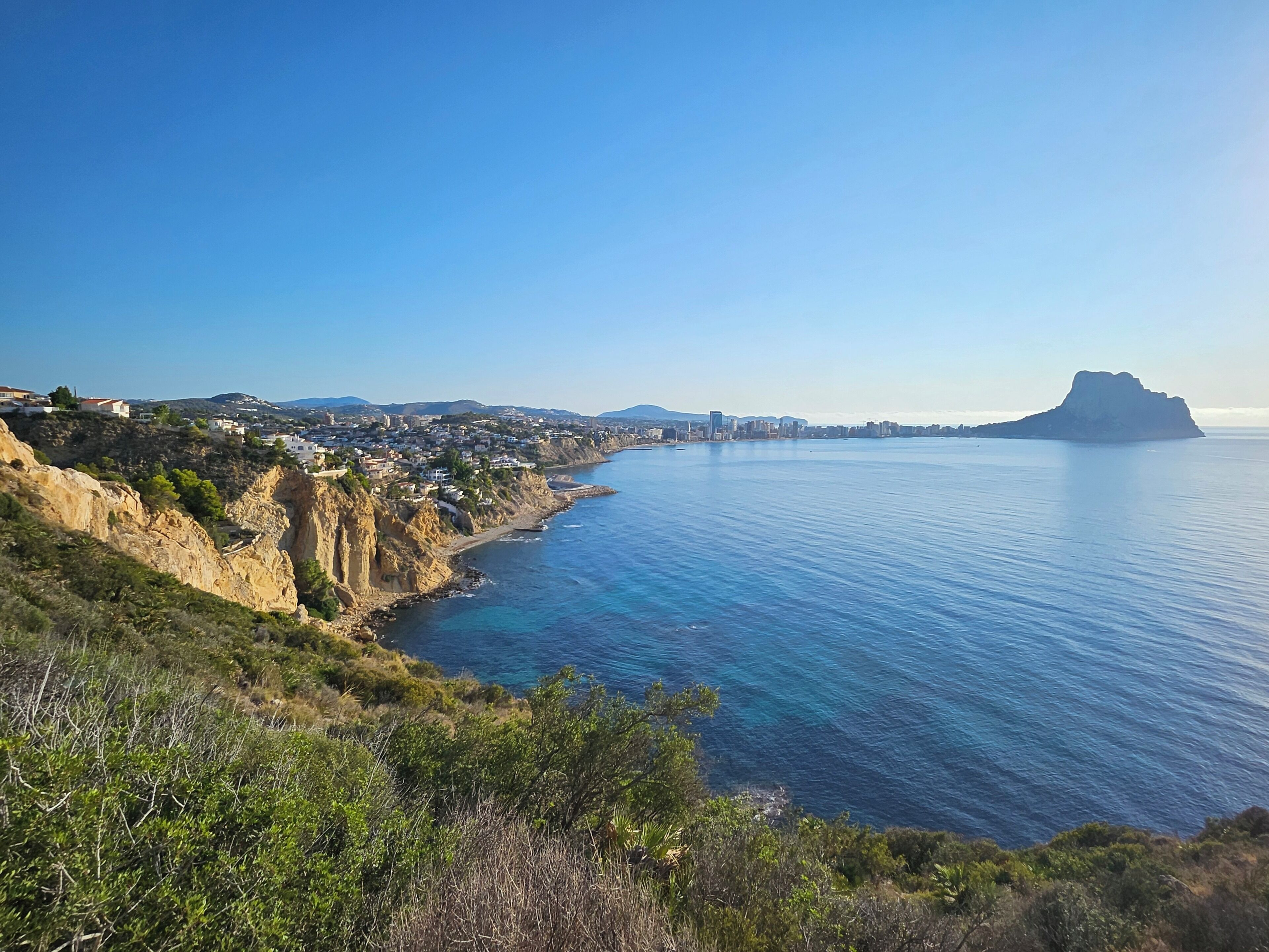 A coastal town with a clear view of the sea, rocky cliffs, and a distant mountain.