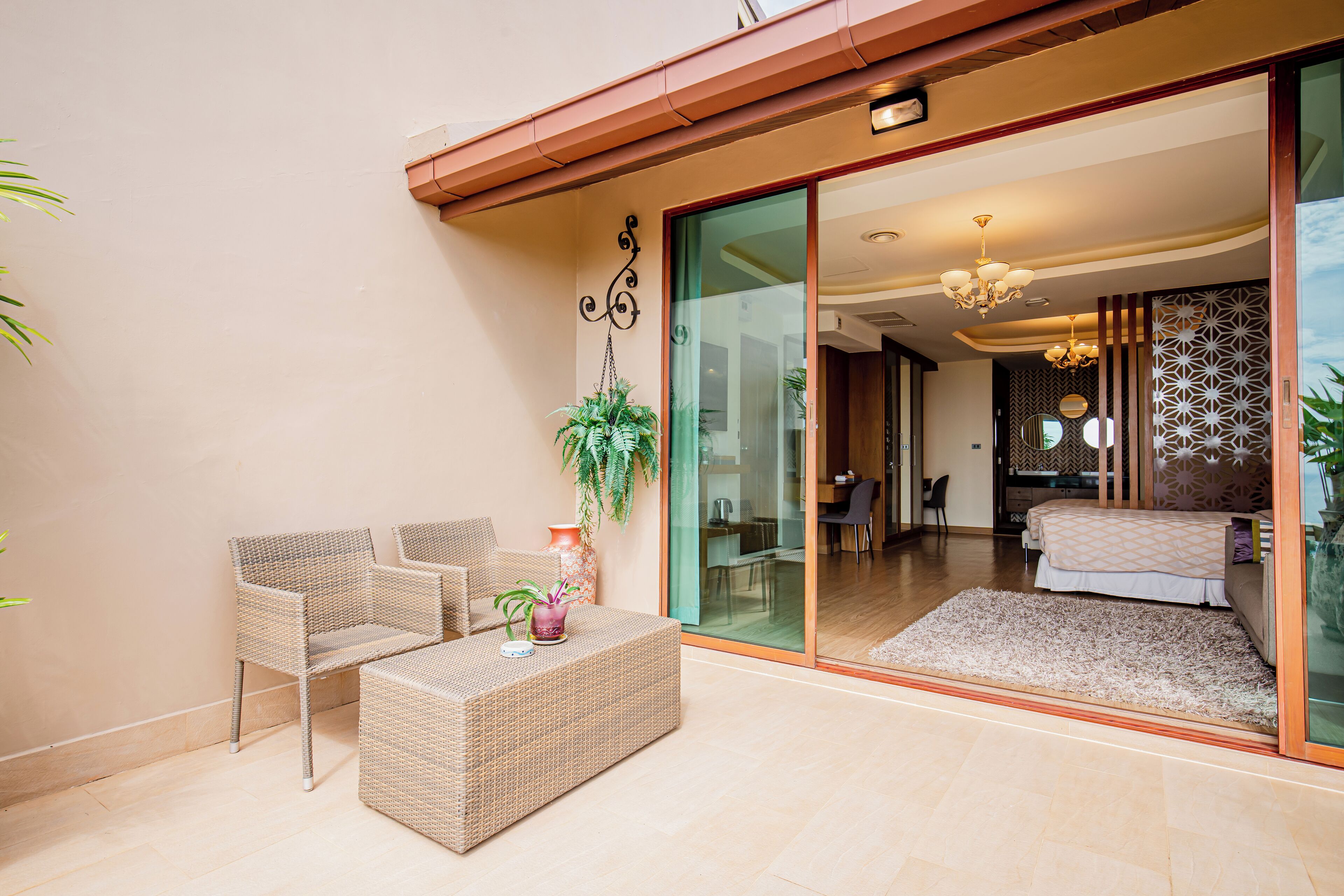 A modern room with a glass door leading to a balcony, wicker furniture, a potted plant, and a bed with a patterned bedspread.