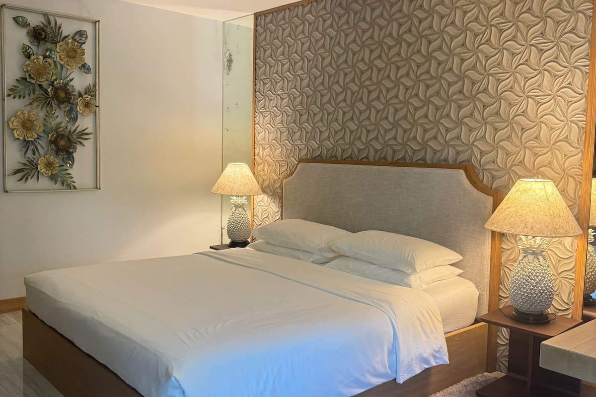 A neatly arranged bedroom with a large bed, two bedside lamps, a decorative wall panel, and a framed floral artwork on the wall.