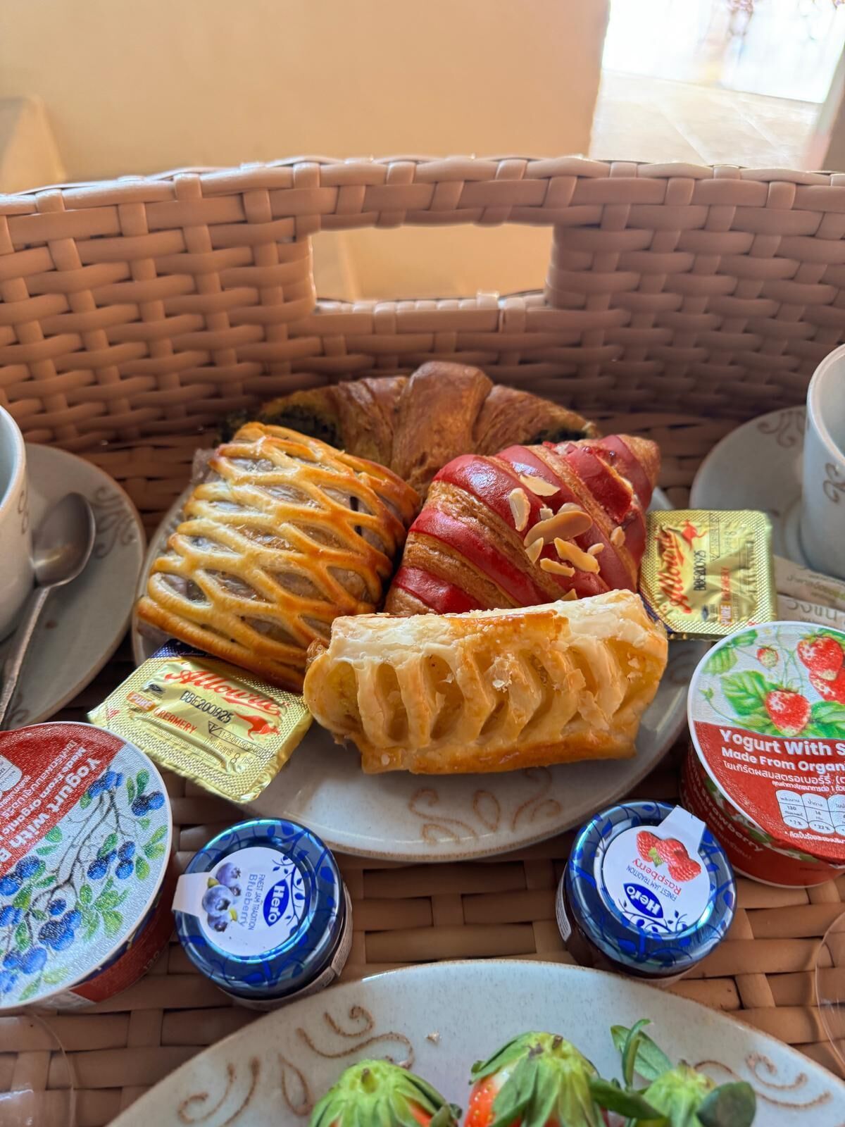 A wicker basket containing pastries, a plate with a pastry, and small containers of yogurt.