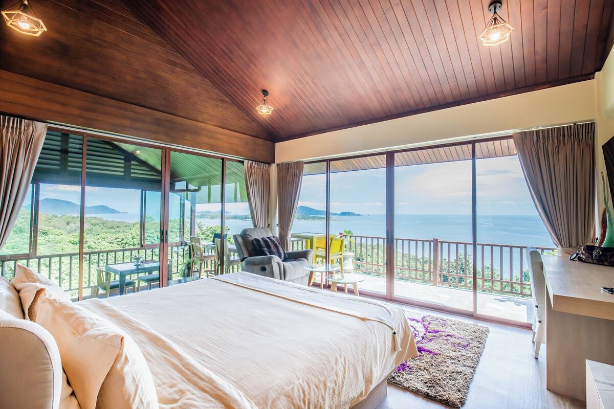 A bedroom with a large bed, a desk, and a balcony with a view of the ocean.