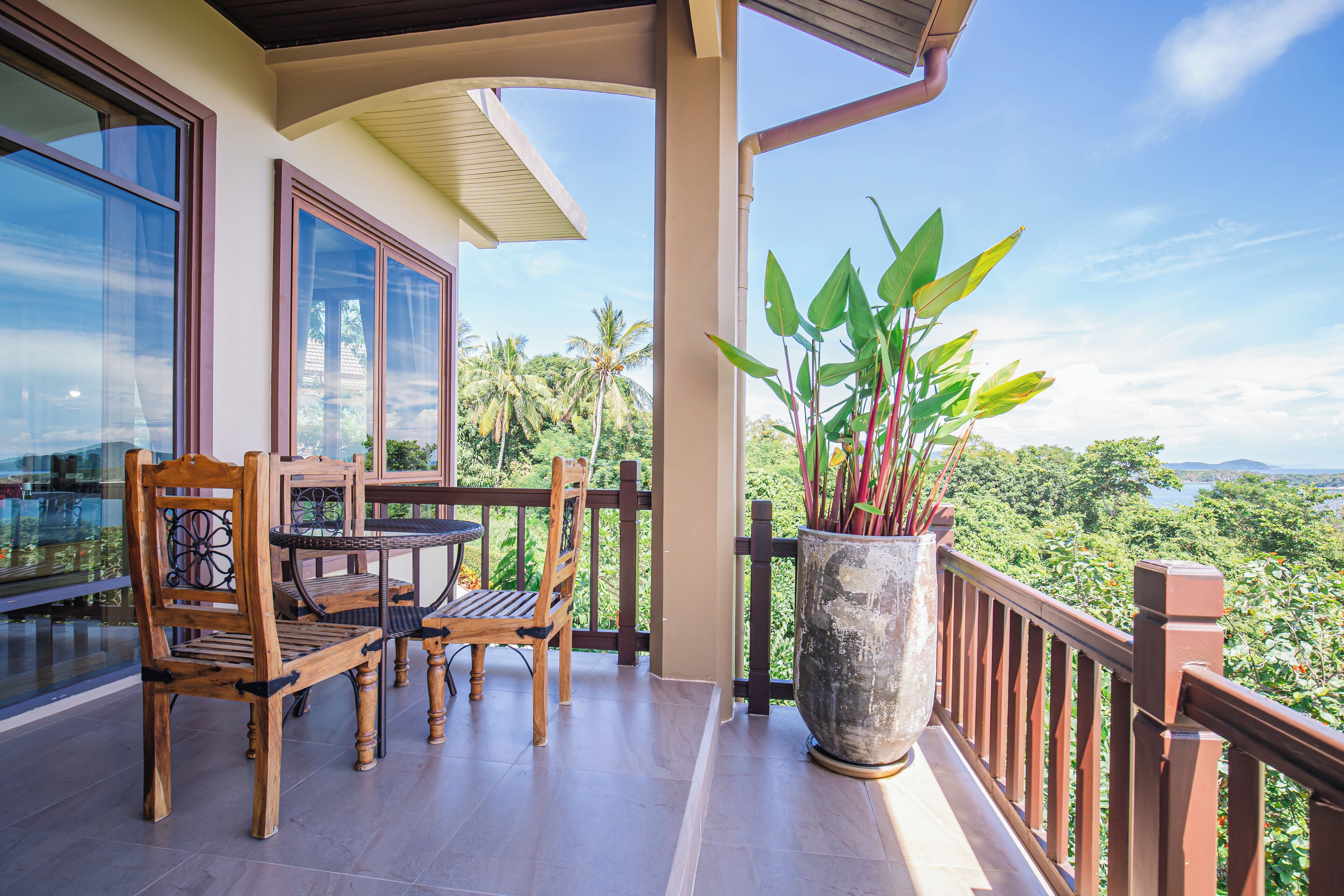 A balcony with wooden furniture, a large potted plant, and a view of a tropical landscape.