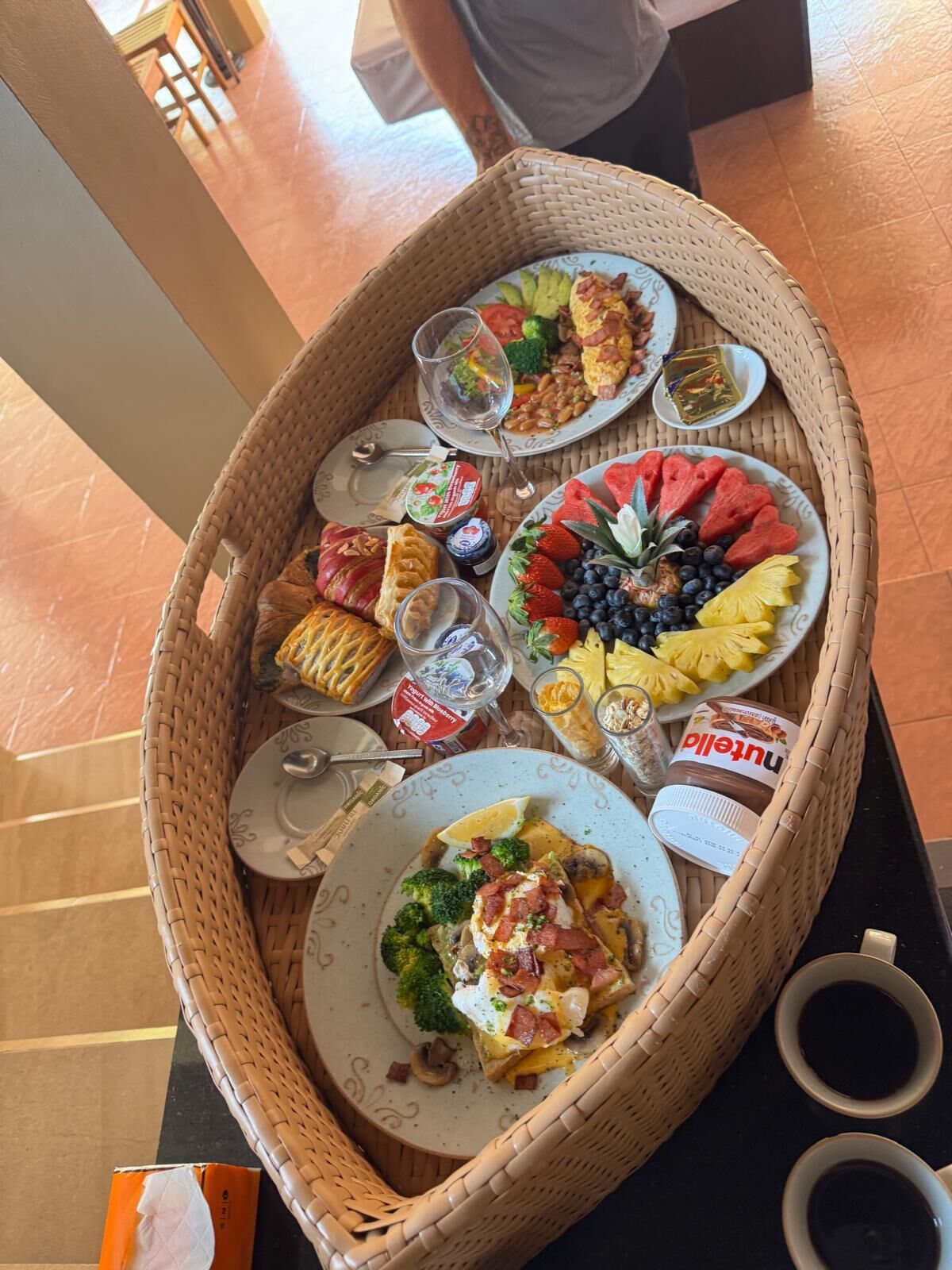 Una cesta de mimbre llena de diversos productos para el desayuno, como bollería, frutas y salsas.