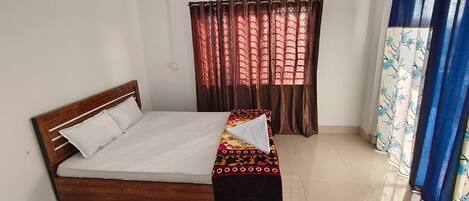 Una cama individual con estructura de madera, colchón blanco y colcha estampada. Se ve una ventana con cortinas marrones y una cortina azul con diseño floral.