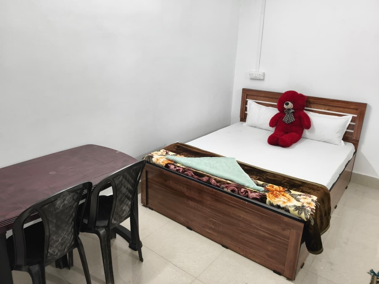 A bedroom with a bed, a wooden headboard, a red teddy bear, a table, and two chairs.