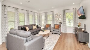 Ein modernes Wohnzimmer mit einem grauen Sofa, zwei Sesseln, einem Couchtisch und einem Fernseher an der Wand.