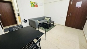 Una habitación pequeña y bien iluminada con un sofá gris, una mesa negra y una silla negra. Tiene una puerta de madera y una ventana pequeña.