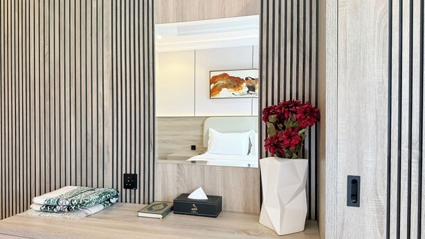 A neatly organized room with a bed, a bedside table with a tissue box and a book, a vase with red flowers, and a framed artwork on the wall.