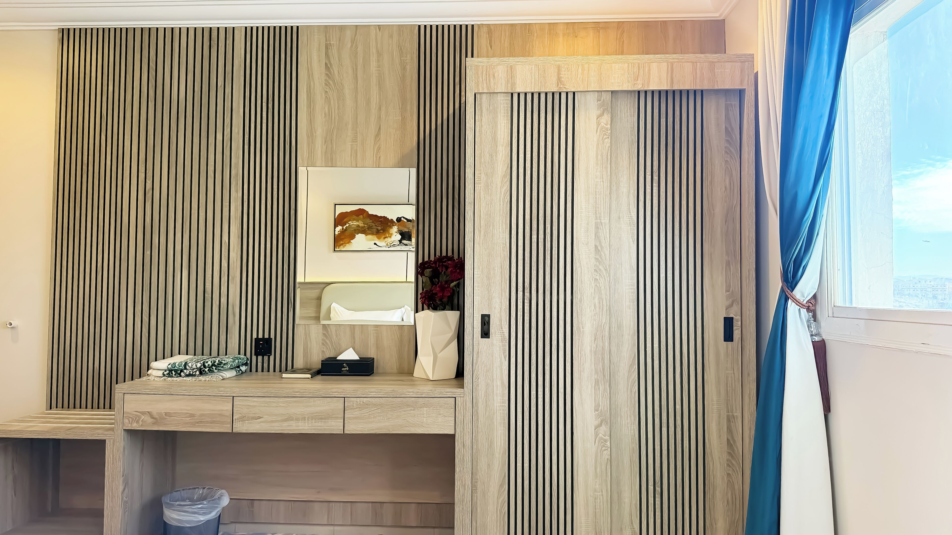 A modern bathroom with vertical wooden paneling, a built-in vanity with a mirror, and a window with blue curtains.